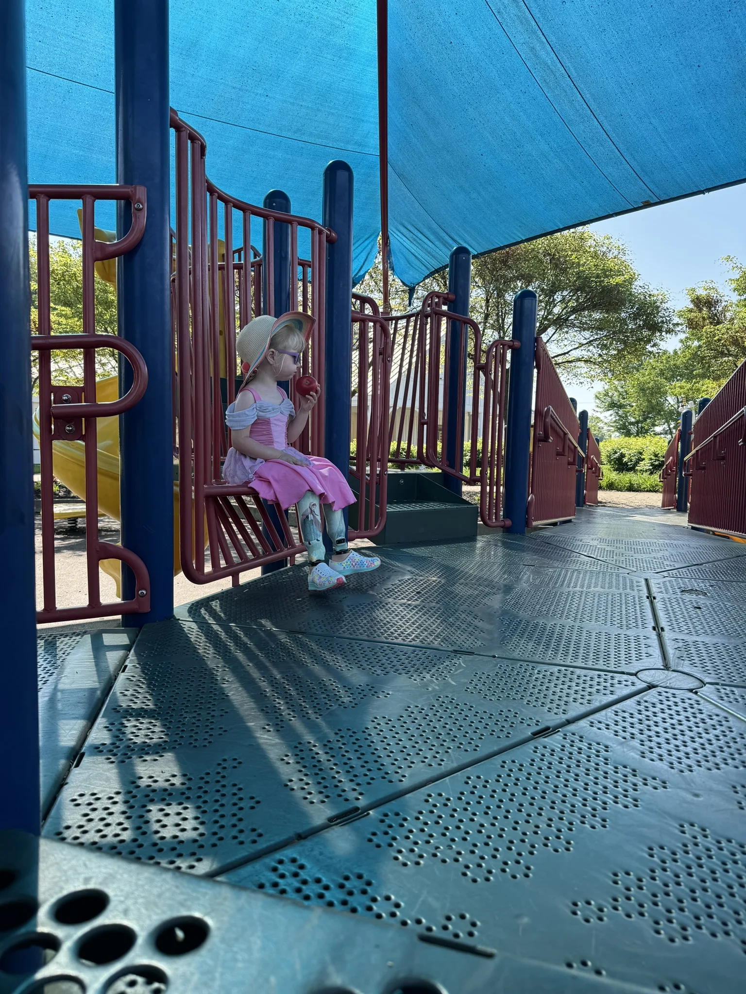 A young girl in a pink princess dress and sun hat sits on an accessible playground ramp at Darree Fields Park, holding an apple.