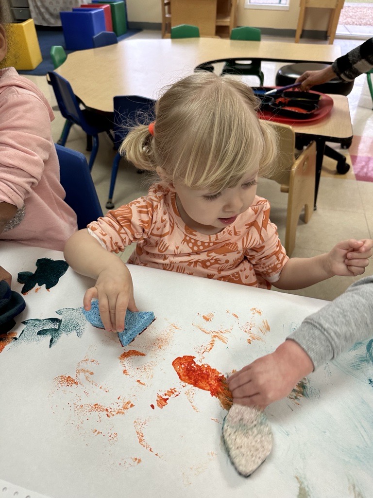 Gwen at Easterseals painting at a shared table with other kids, using foam shapes and bright colors