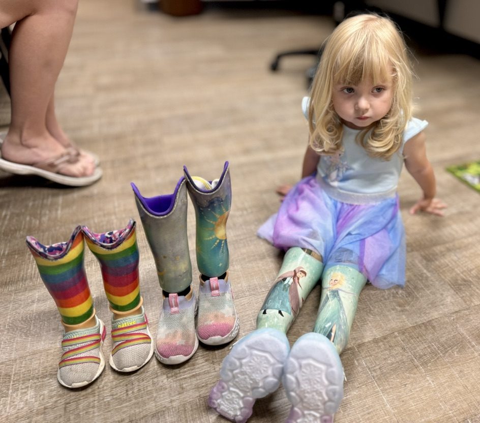 Gwen sitting on the floor with her first, second, and third sets of prosthetics lined up to show how she has grown